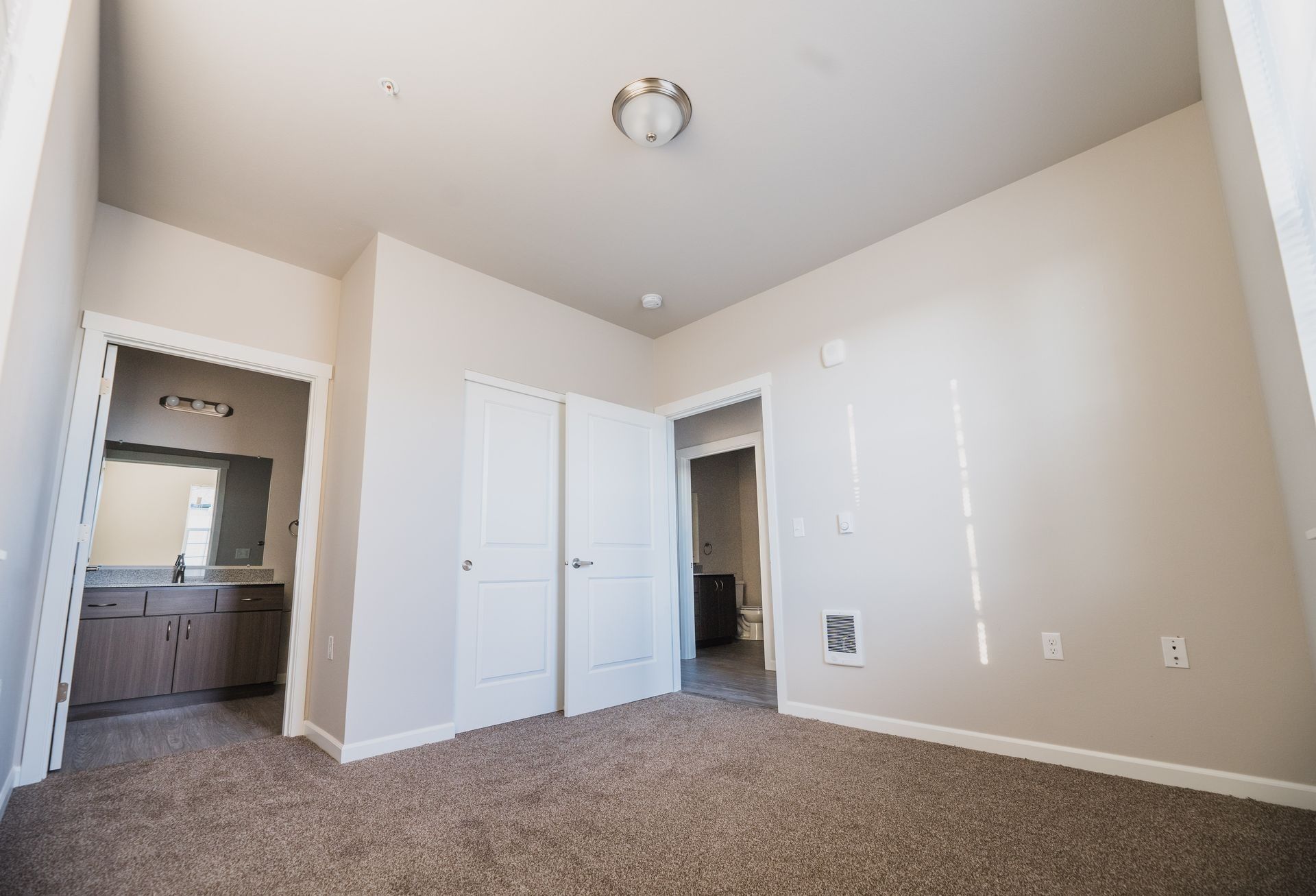 Empty beige bedroom with carpet, white closet doors, and an open doorway to a bathroom.