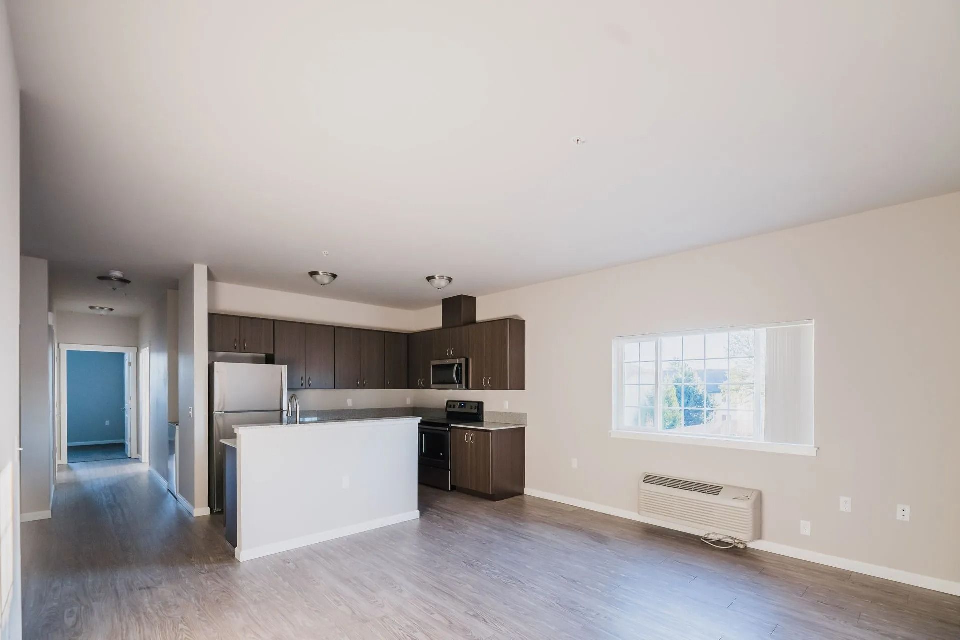 Empty living room with open kitchen, light wood floors, white walls, and a window with radiator beneath it