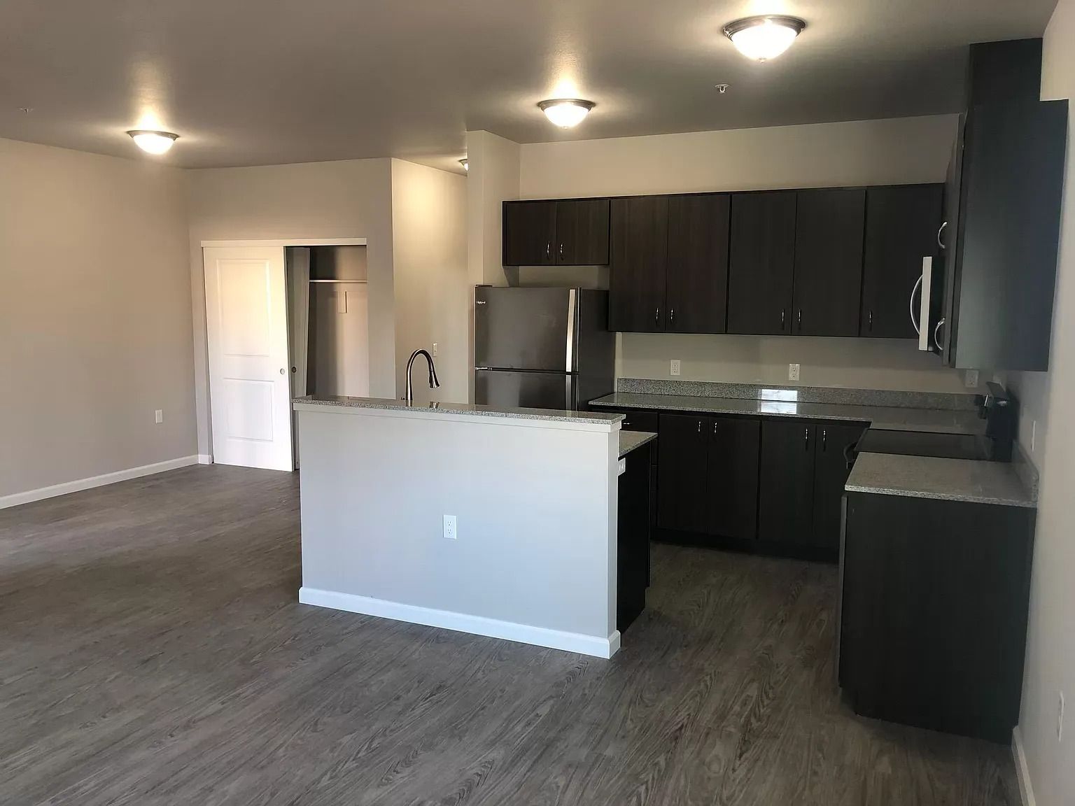 Empty modern kitchen and living area with gray carpet, dark cabinets, and white breakfast bar.