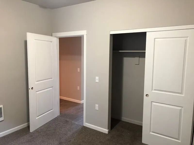 Empty bedroom with beige walls, gray carpet, and two open white doors, one to a hall and one to a closet