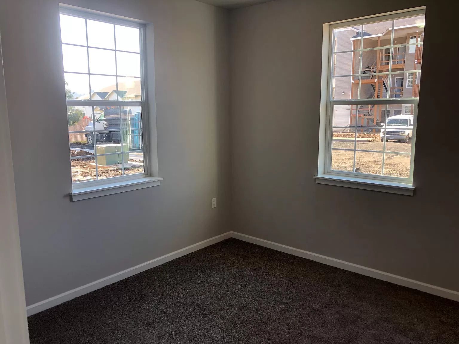 Empty carpeted room with two windows and gray walls, lit by daylight.