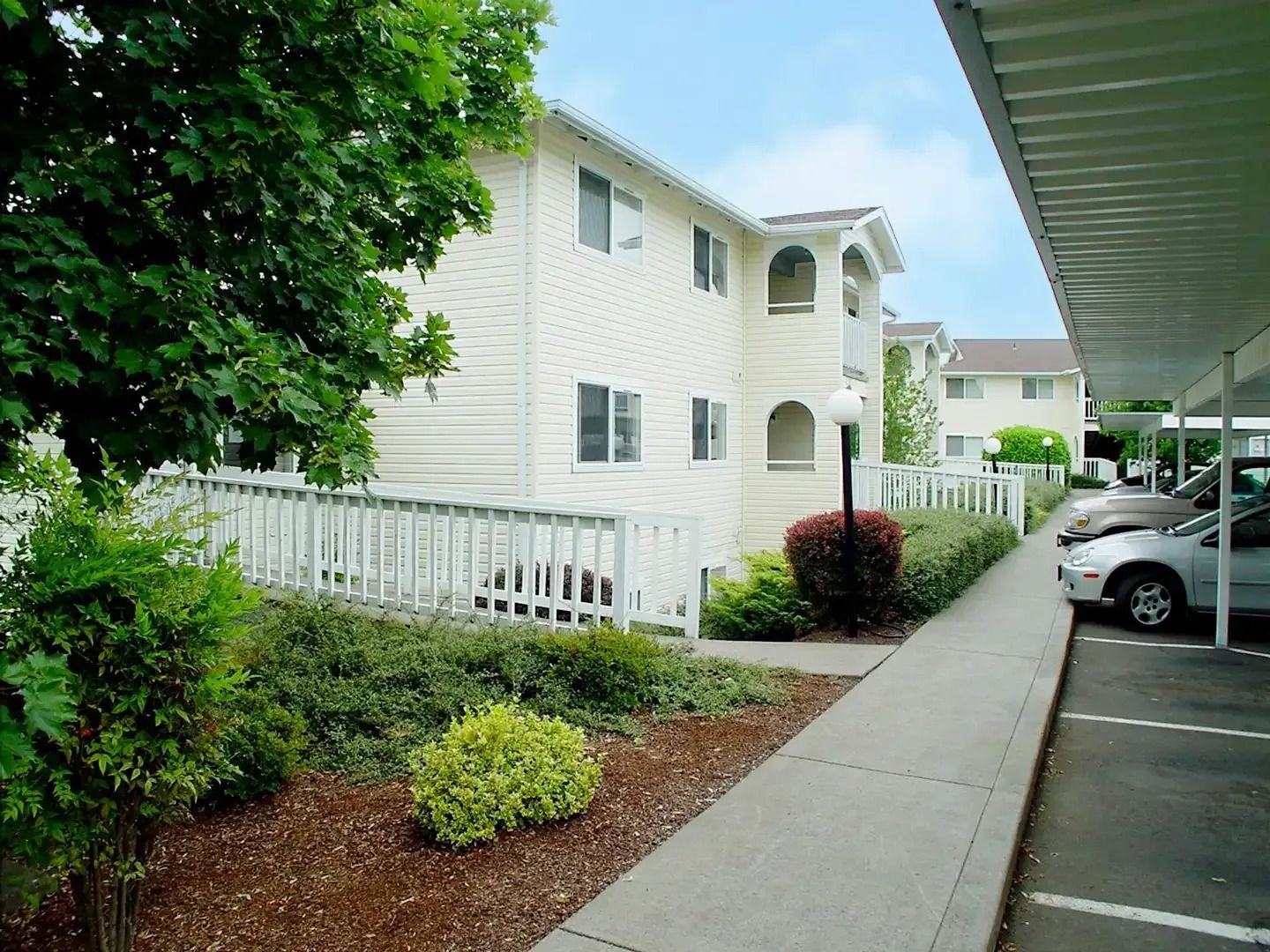 Apartment walkway beside white buildings, with landscaping and parked cars under a covered lot.