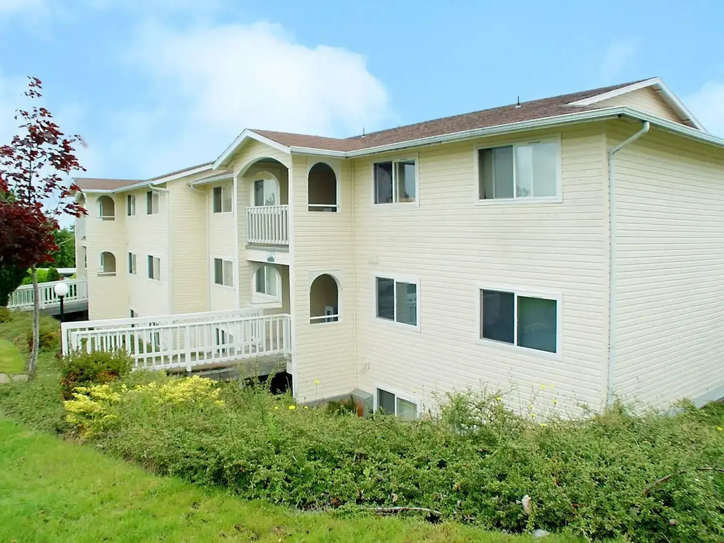 Cream-colored apartment building with balconies and windows, set beside green shrubs under a blue sky