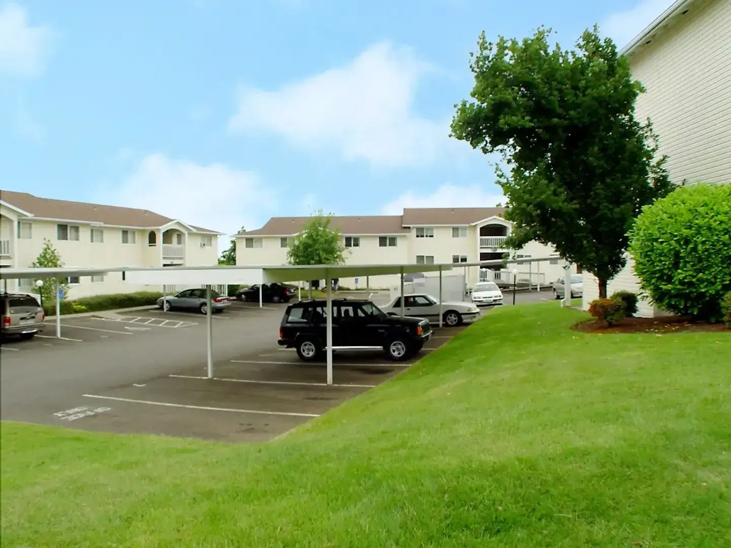 Apartment parking lot with parked cars, white townhouses, and a grassy slope under a blue sky