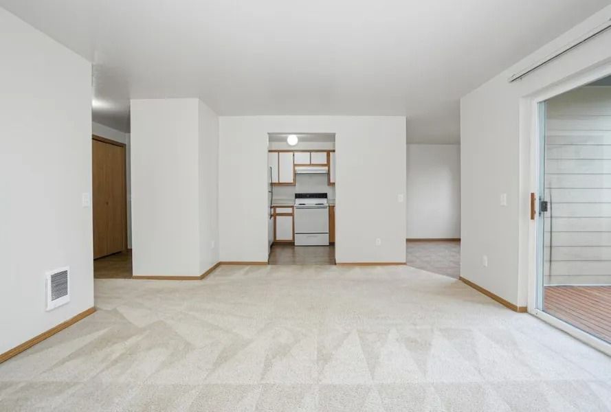 Empty beige living room with carpet, white walls, and a kitchen pass-through in the center