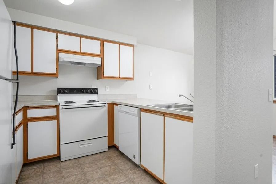 Small white kitchen with wood-trim cabinets, stove, sink, and dishwasher in an empty room
