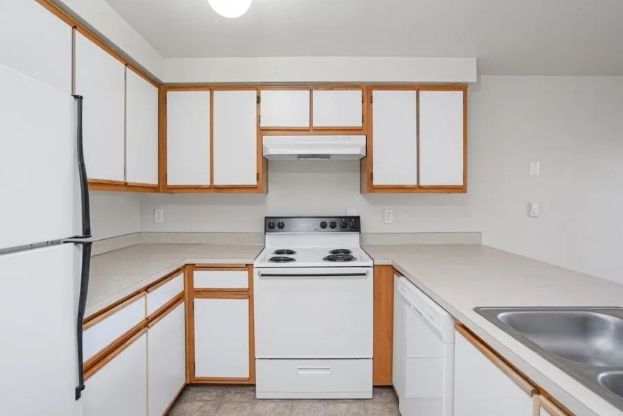 Small white kitchen with wood-trim cabinets, stove, refrigerator, and sink on the right.