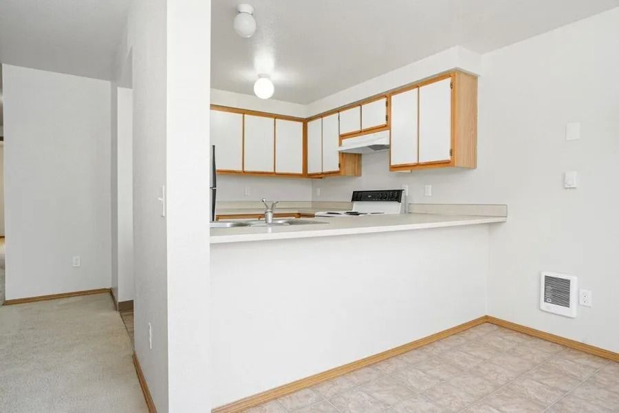 Empty apartment kitchen with white cabinets, light countertops, and beige carpeted living area.
