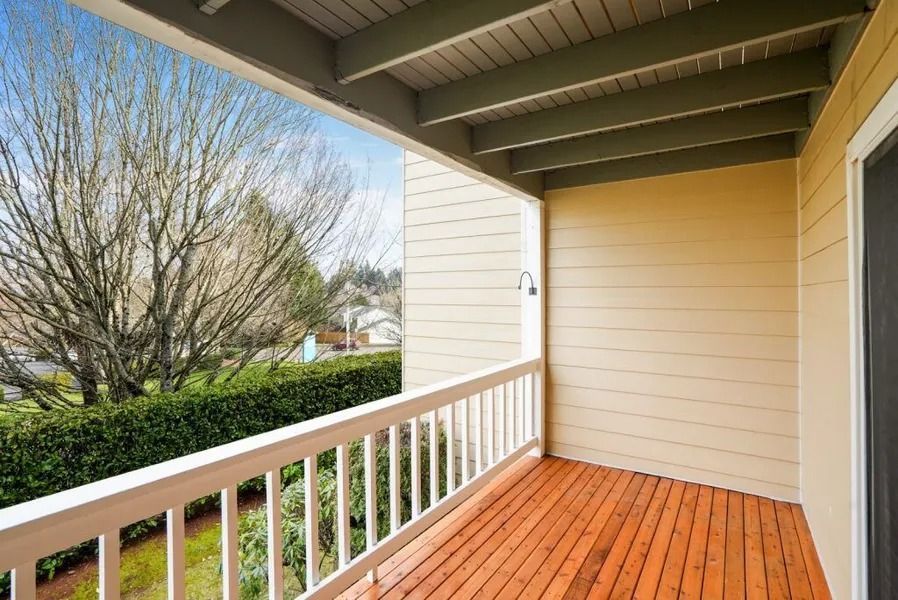 Covered front porch with wooden floor, white railing, beige siding, and view of trees and hedges.