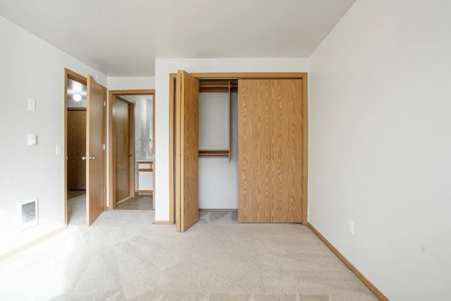 Empty beige bedroom with open closet and bathroom doorway, white walls, and carpeted floor