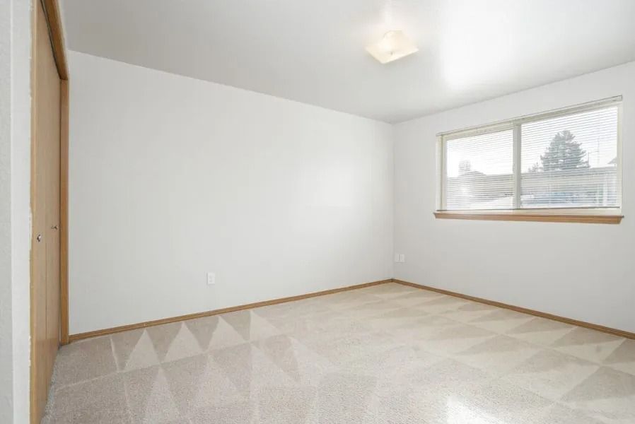 Empty white bedroom with beige carpet, wood trim, and a window with blinds.