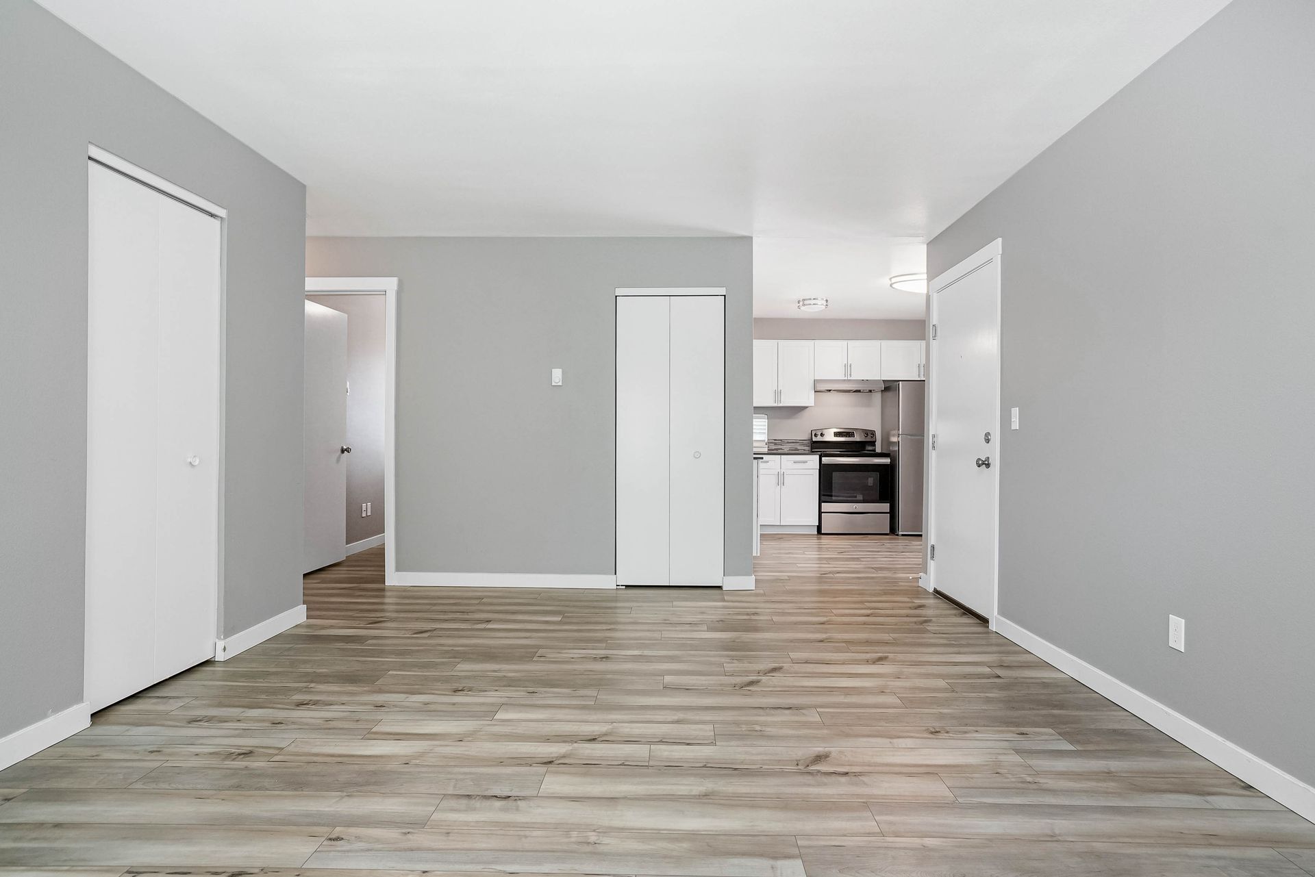 Empty gray living room with wood floors, open doorway, and a view into a small kitchen.