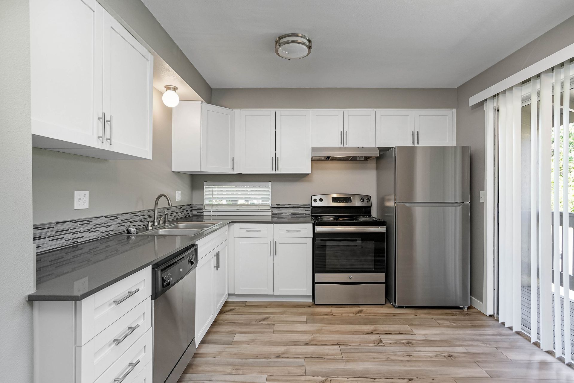 Bright modern kitchen with white cabinets, gray counters, stainless appliances, and sliding glass doors.