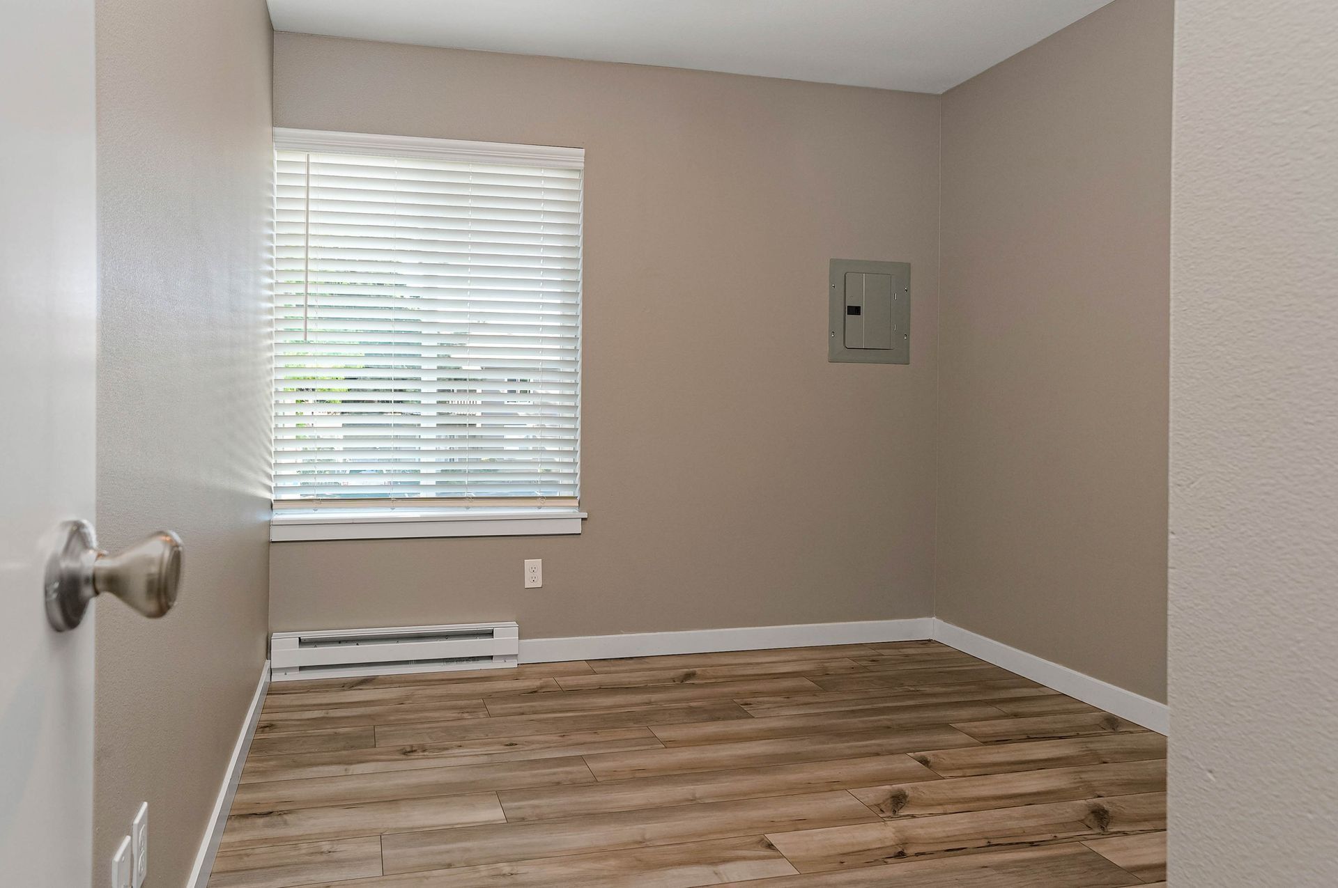 Empty beige room with window blinds, white trim, and wood-patterned floor.