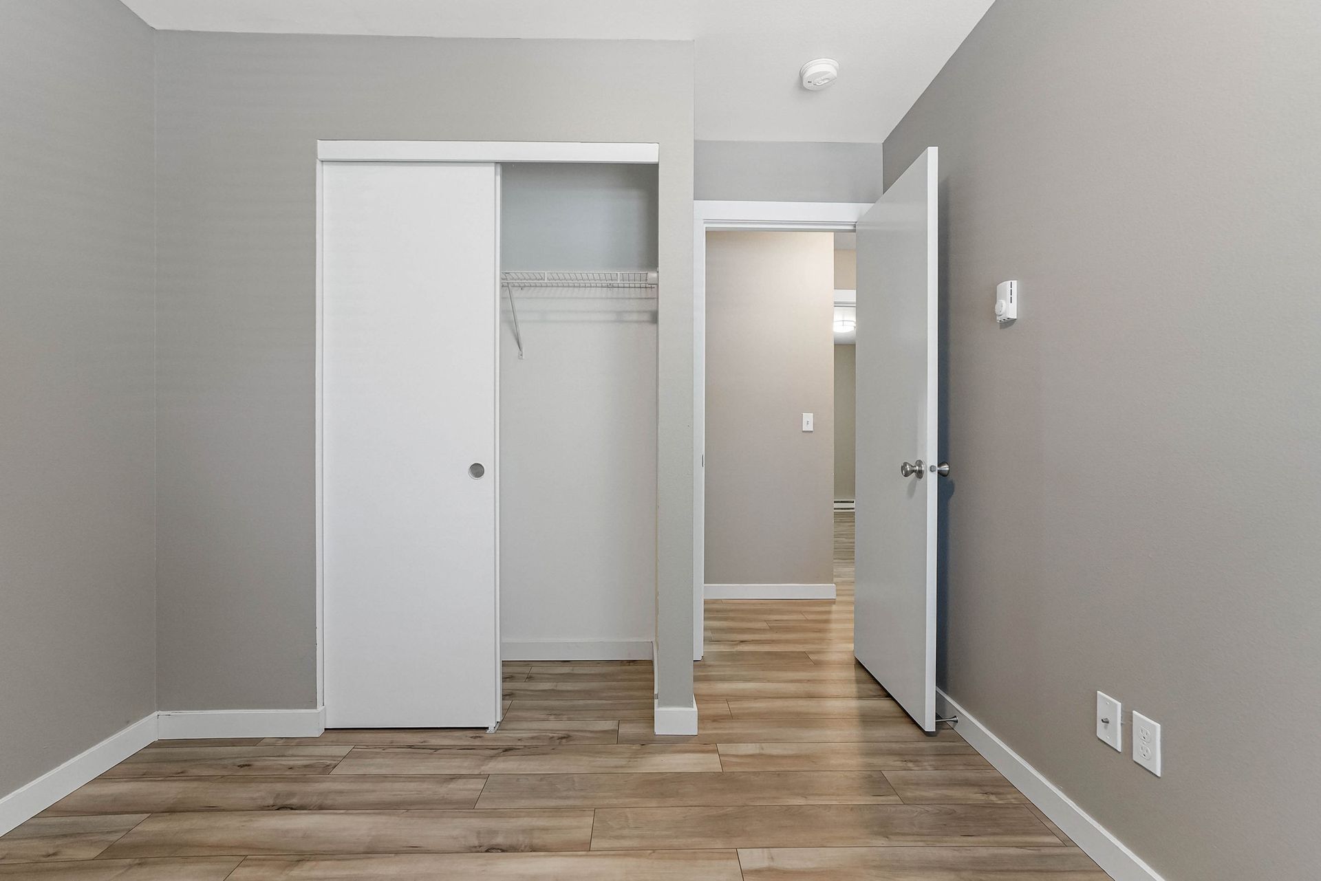 Empty gray bedroom with open closet, wood floor, and open door to hallway