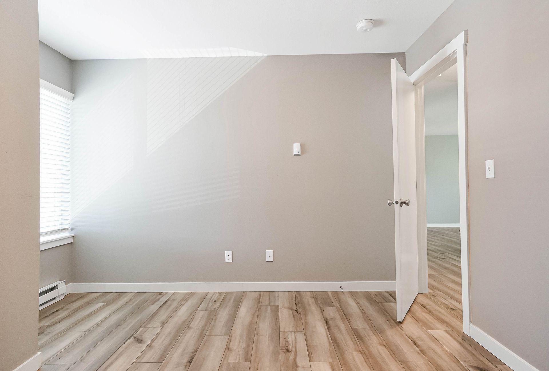 Empty gray bedroom with wood floors, a window on the left, and an open door to another room.