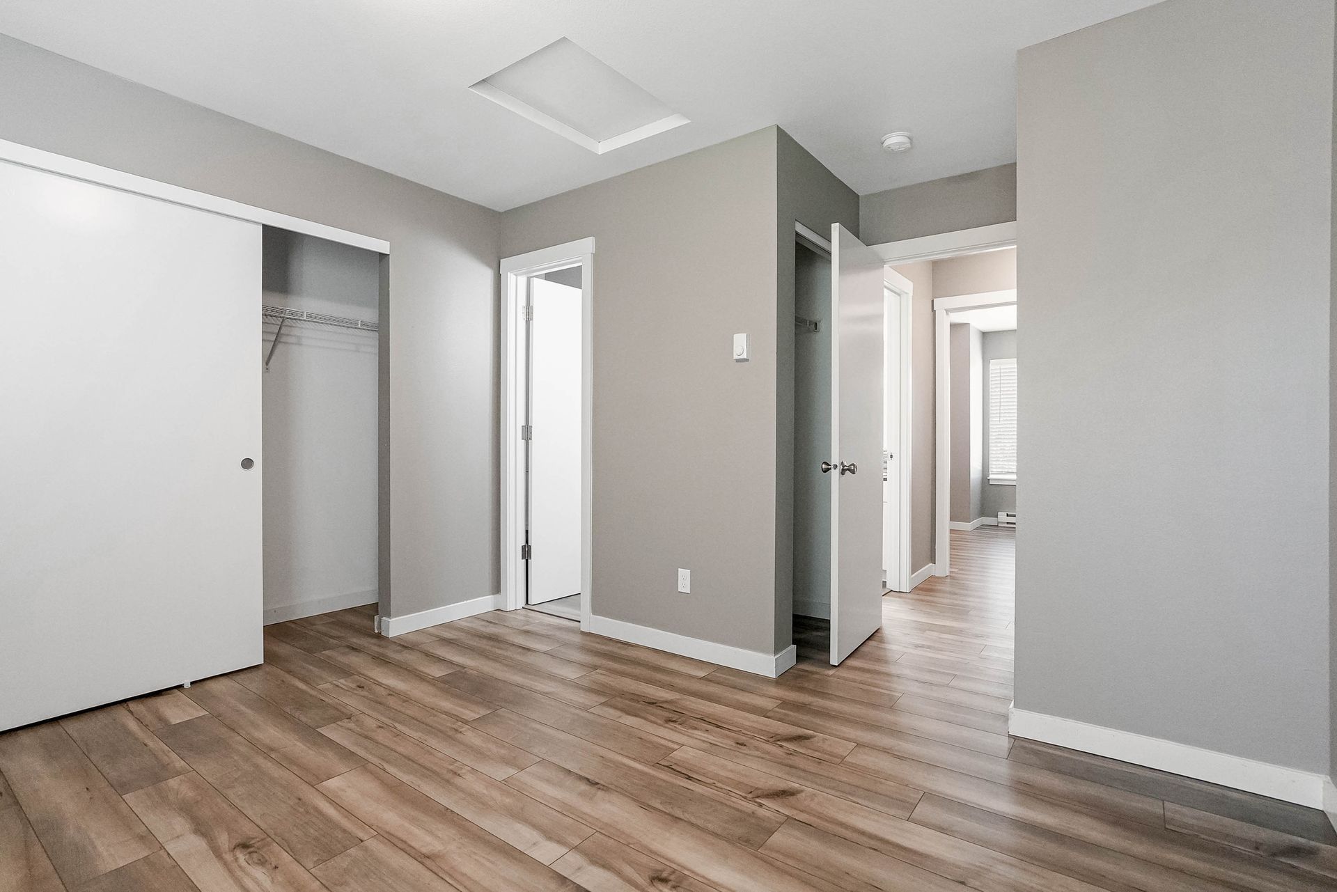 Empty modern room with gray walls, wood floor, open closets, and a hallway into another room