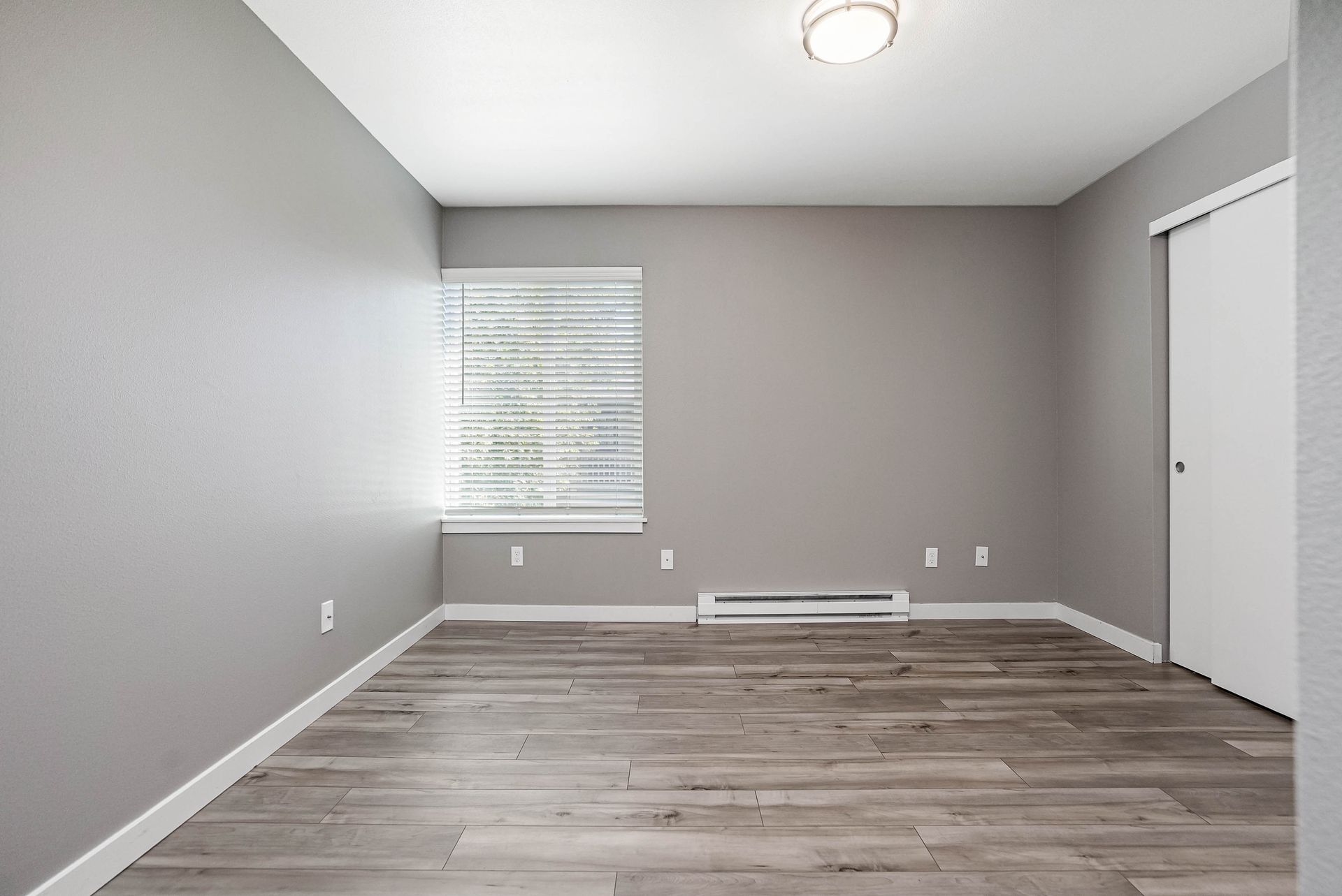Empty gray bedroom with wood-look flooring, window blinds, and a closed white closet door.
