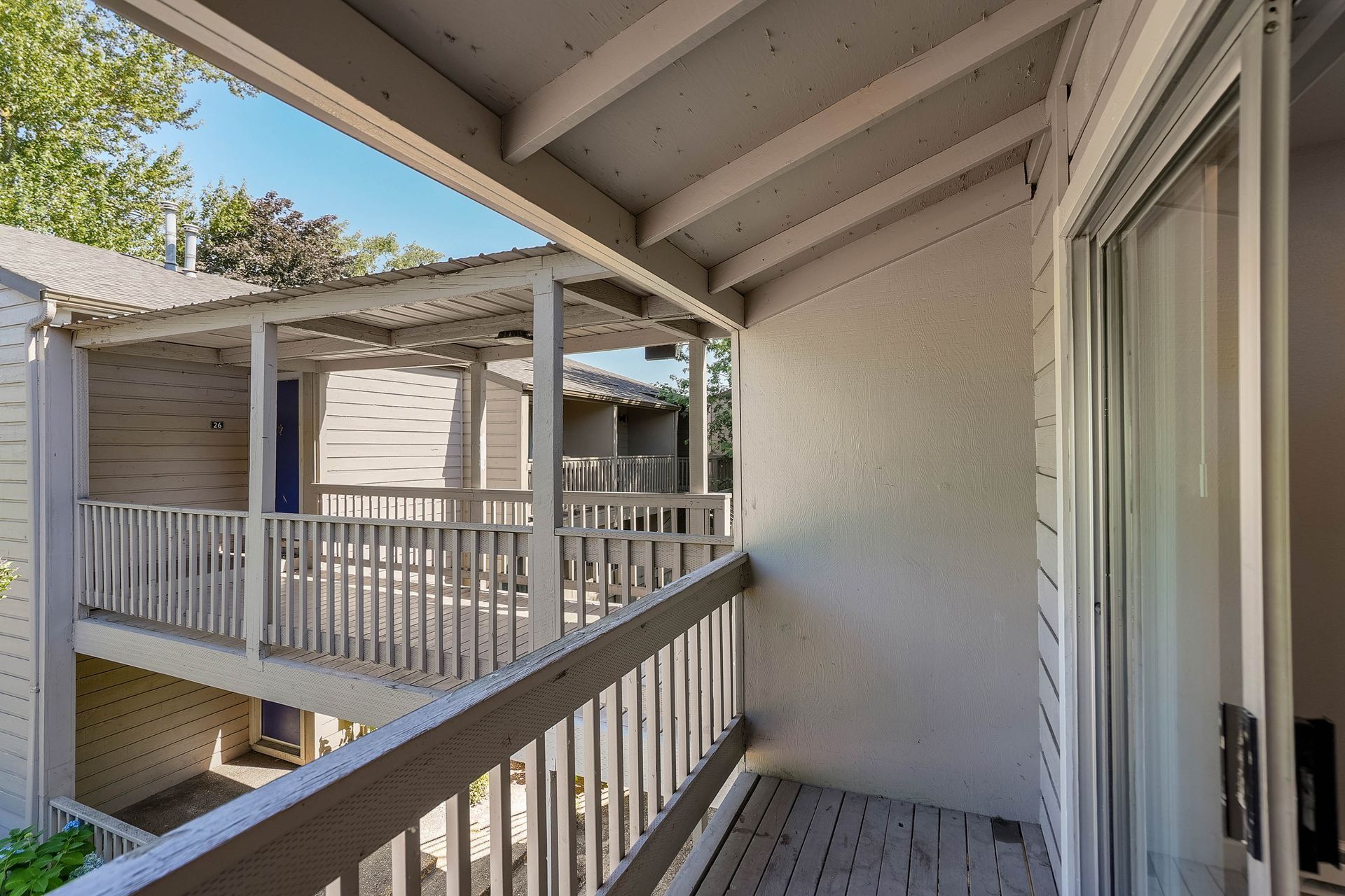 Apartment balcony with white railing and sliding glass door, overlooking neighboring unit and blue sky