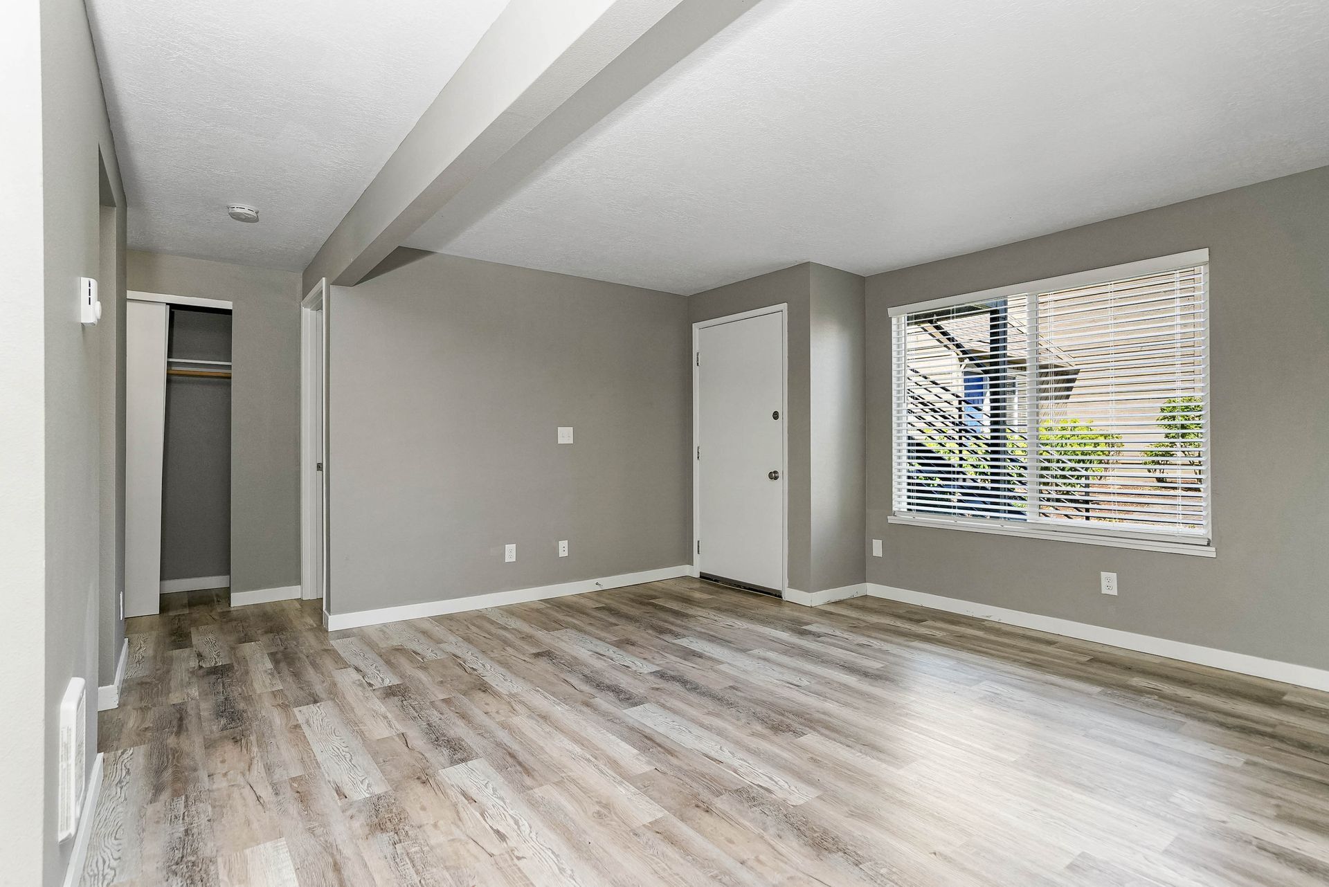 Empty gray living room with wood-look flooring, a window with blinds, and a hallway doorway
