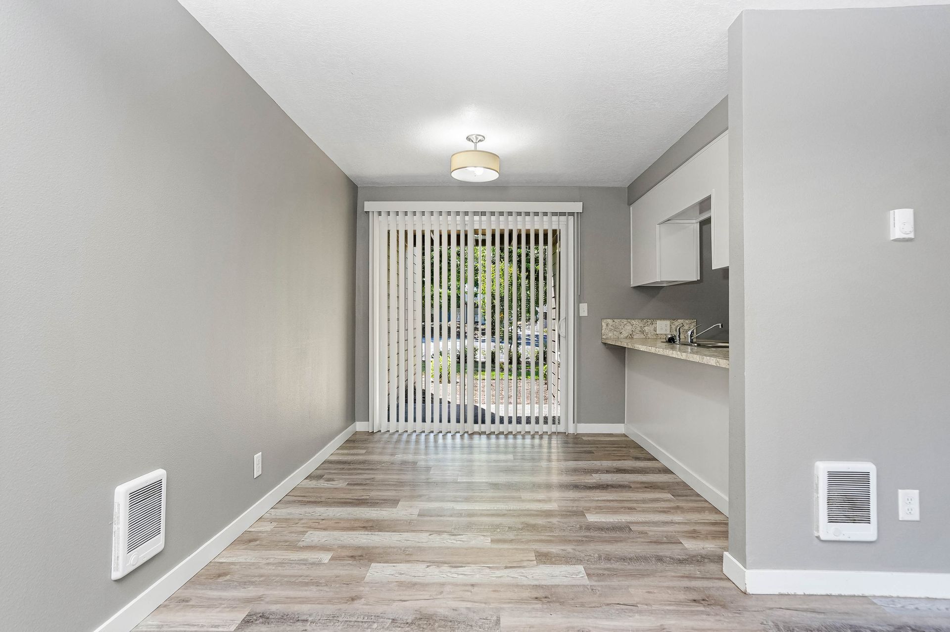 Empty modern living room with gray walls, wood floors, kitchen pass-through, and sliding glass door with vertical blinds