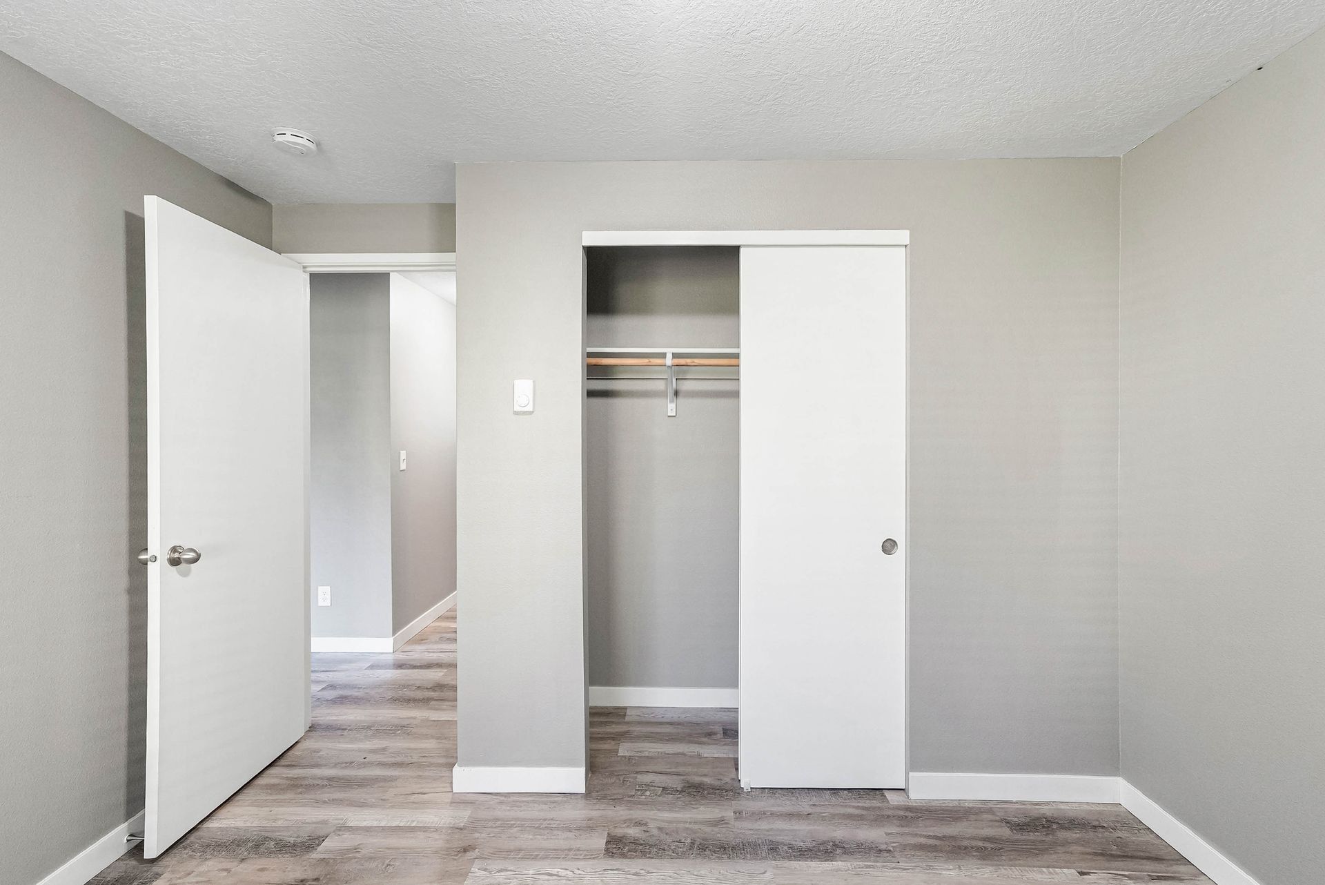 Empty bedroom with open closet and hallway doorway, white walls and gray wood-look flooring.