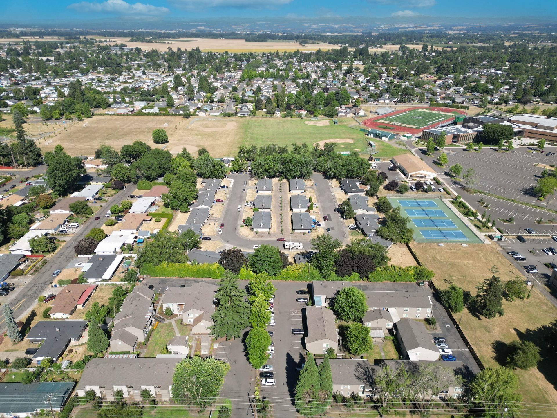 Aerial view of a suburban neighborhood with homes, streets, trees, and a sports field in the distance