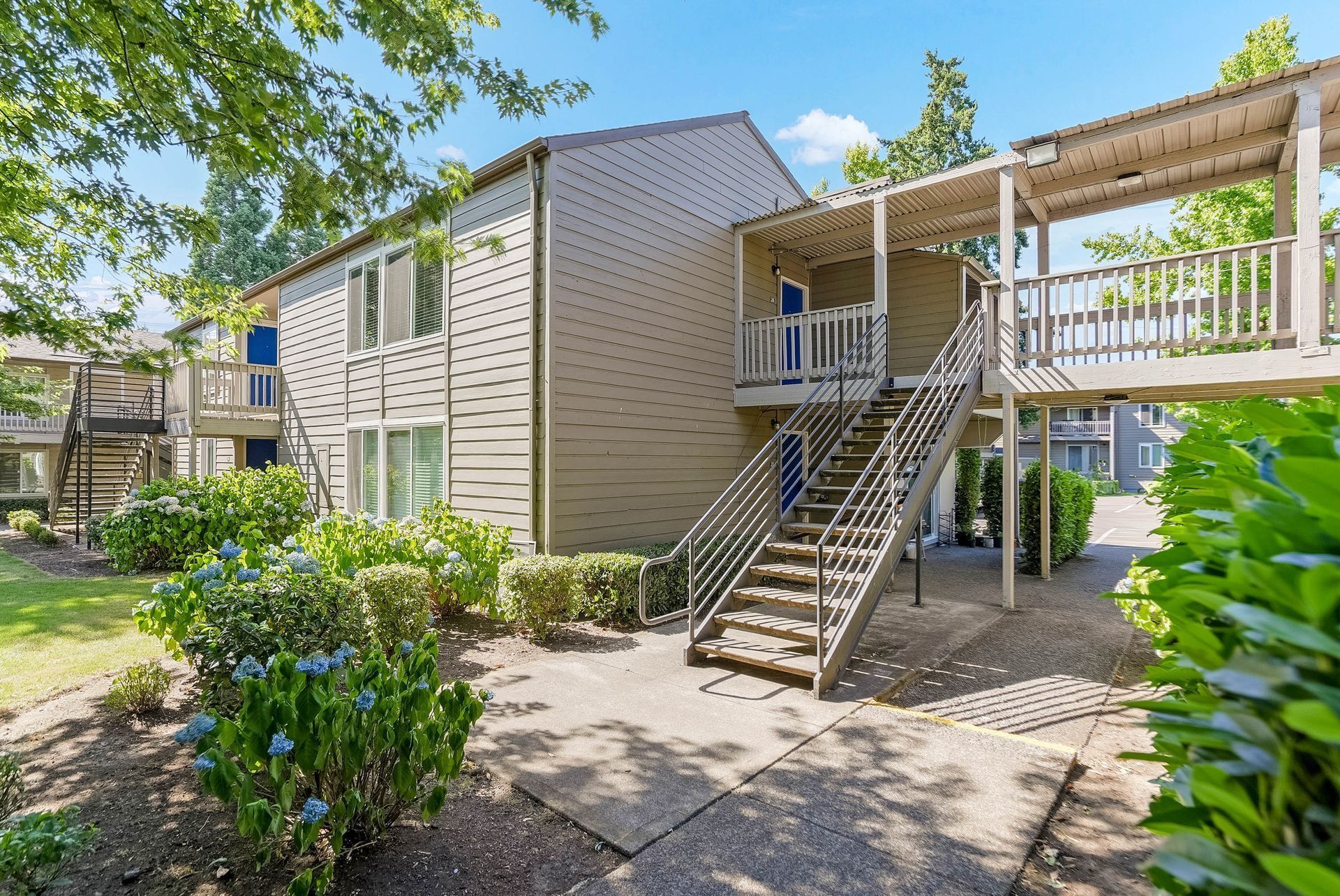 Apartment exterior with stairs, balconies, and green landscaping on a sunny day
