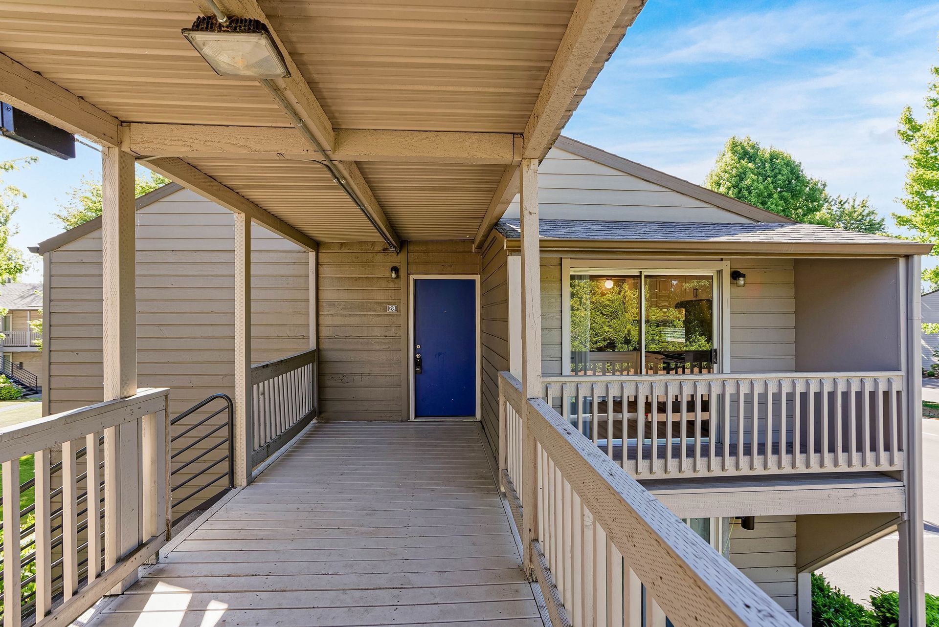 Covered wooden walkway leading to a beige apartment entrance with a blue door and balcony railings