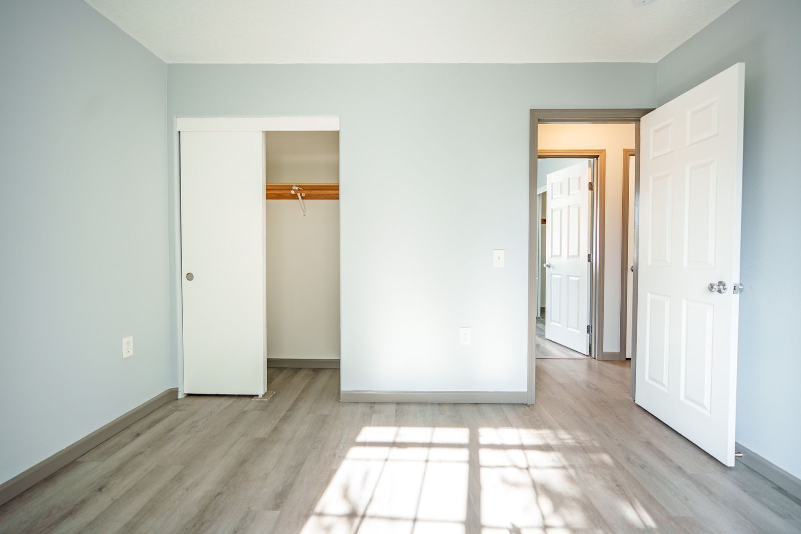 Empty bedroom with light gray walls, wood flooring, open closet, and sunlight through a window.