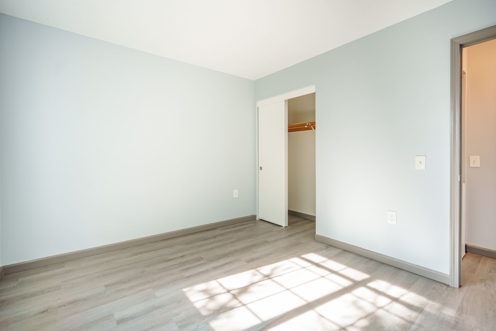 Empty light blue bedroom with gray wood floors, open closet door, and sunlight on the floor.