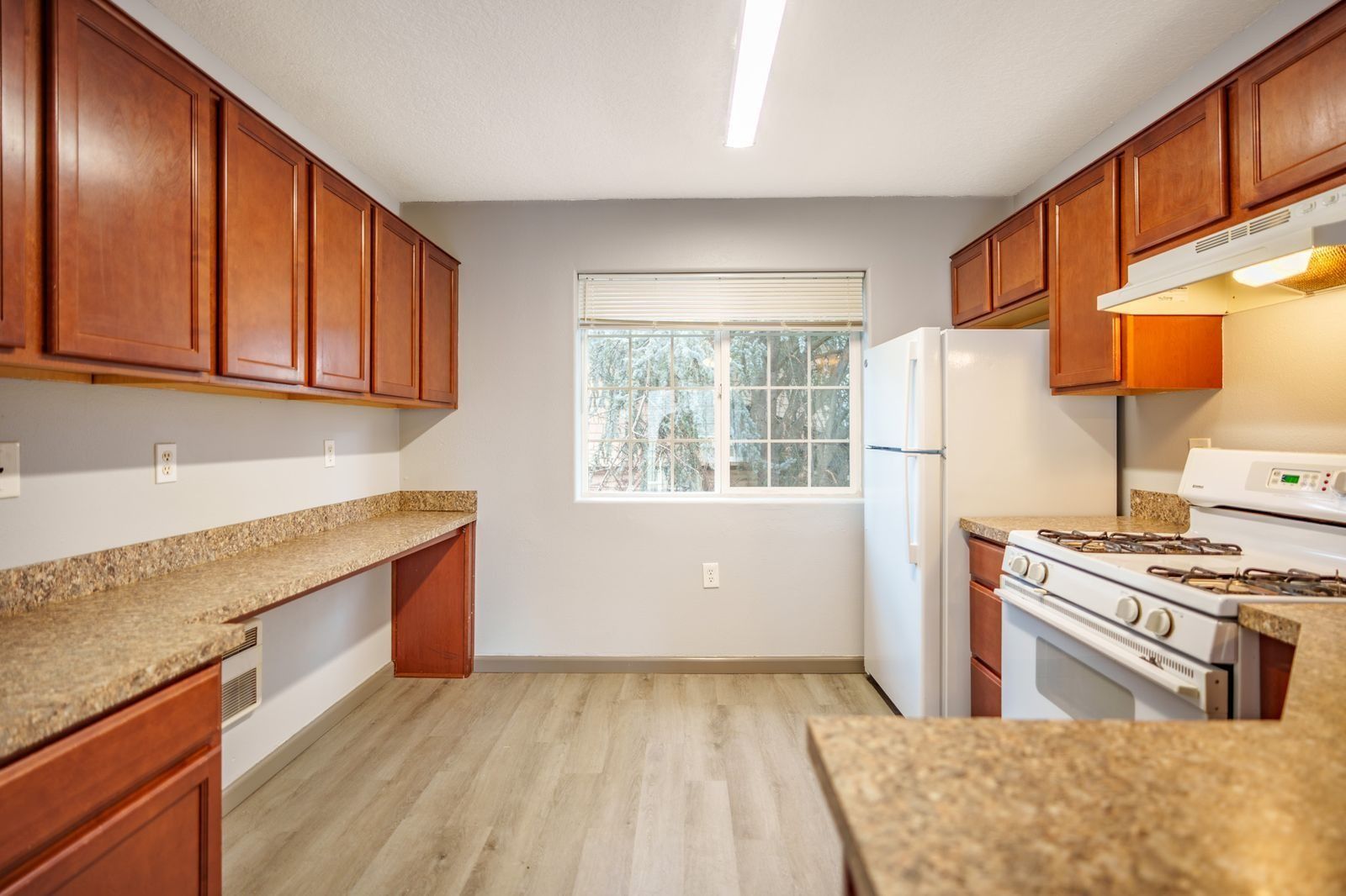 Empty kitchen with wood cabinets, granite counters, white appliances, and a window over wood-look flooring