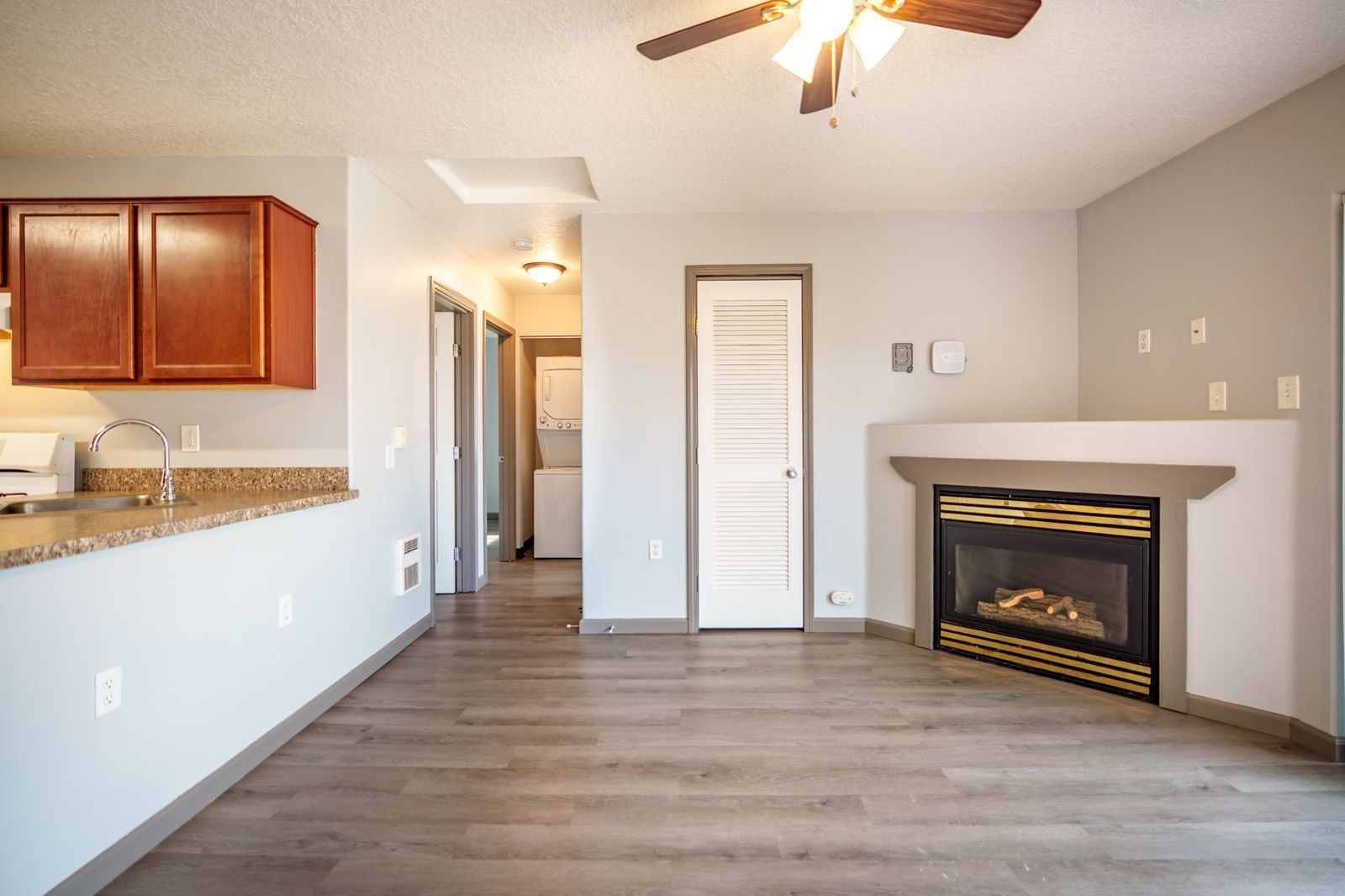 Empty living room with gray walls, wood floors, ceiling fan, fireplace, and kitchen pass-through on the left