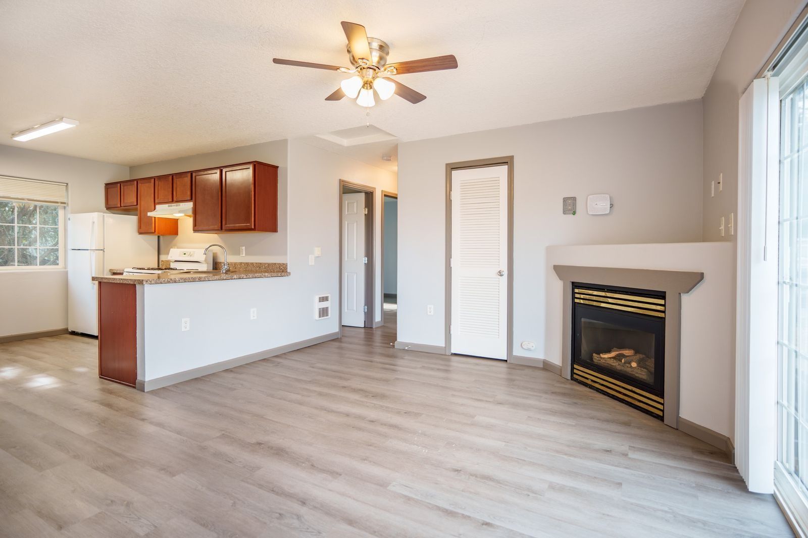 Empty living room with kitchen pass-through, ceiling fan, fireplace, and light wood flooring