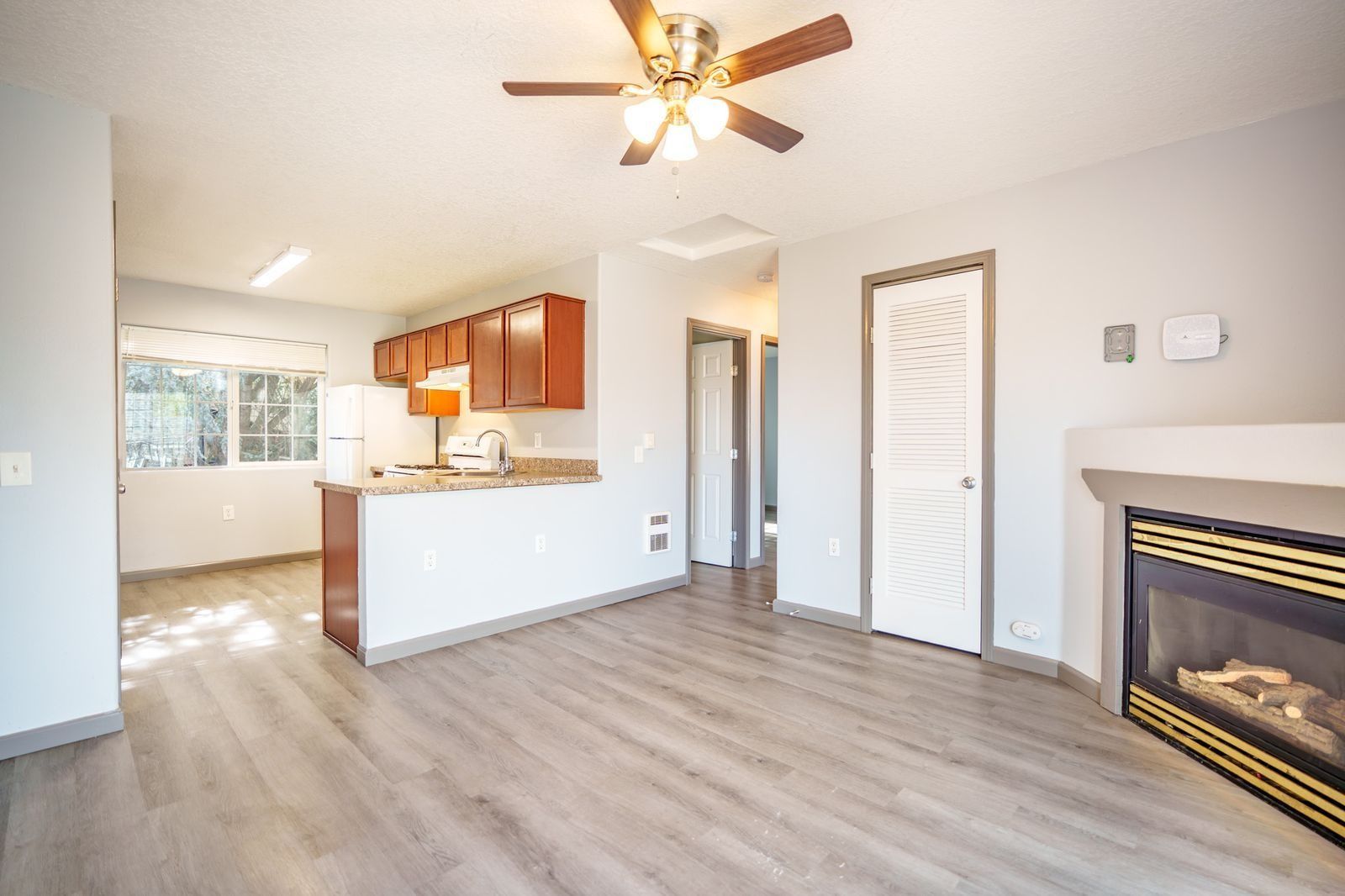 Empty living room with ceiling fan, fireplace, wood floor, and open kitchen area