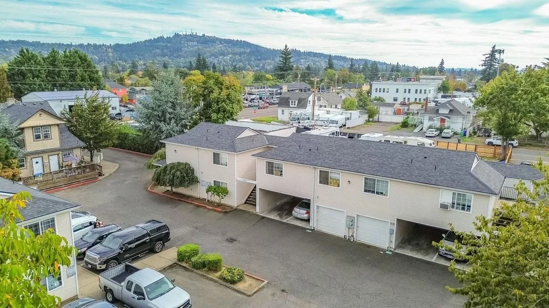 Aerial view of a white apartment complex with parked cars, trees, and hills in the background