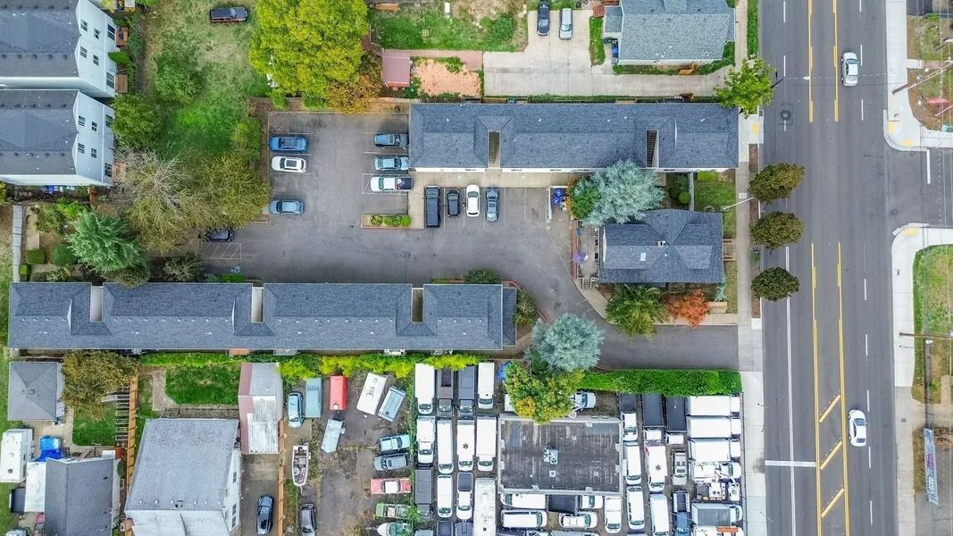 Aerial view of a parking lot and buildings beside a road, with many parked vehicles and trees.