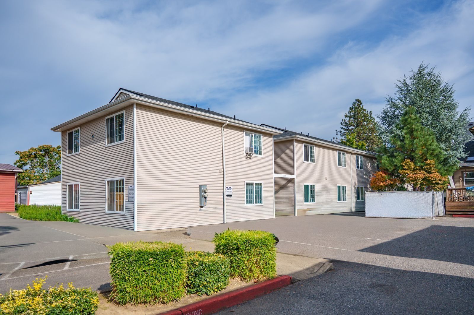 Two-story beige apartment building with parked cars and trimmed hedges under a blue sky