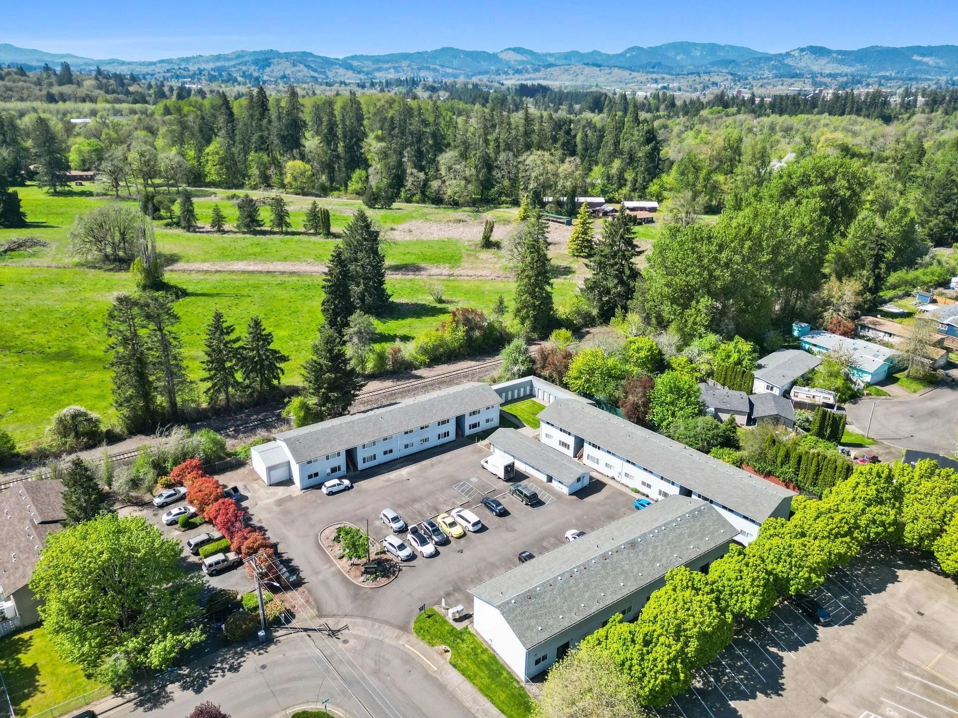 Aerial view of a white building complex with parked cars, surrounded by green fields and trees.