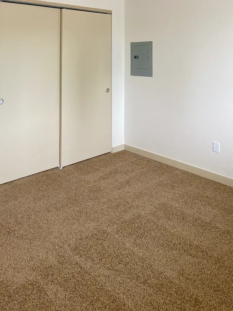 Empty beige-carpeted room with closed sliding closet doors and a wall-mounted electrical panel.