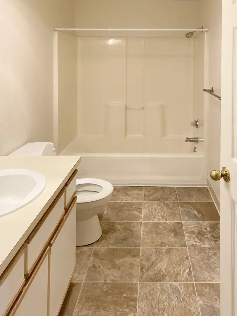 Empty beige bathroom with a tiled floor, sink vanity, toilet, and bathtub with sliding glass doors.