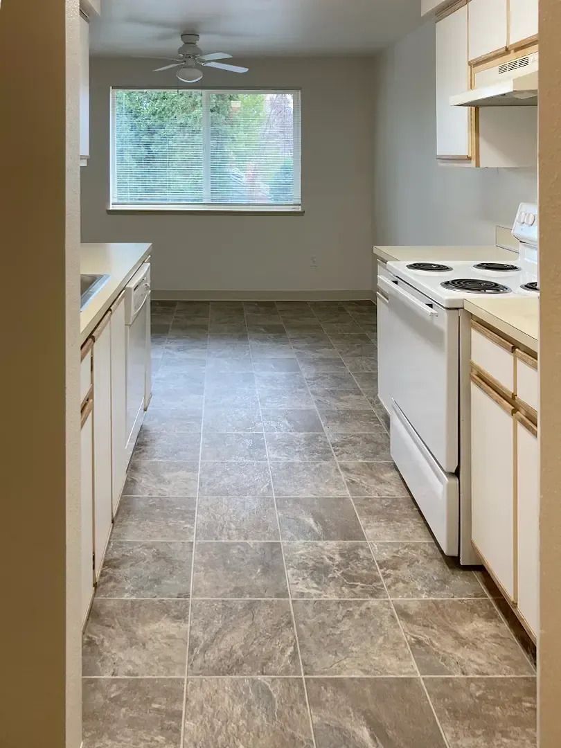 Empty galley kitchen with gray tile floor, white cabinets, and a window at the far end.