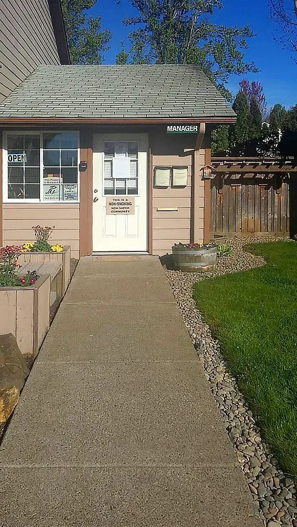 Front walkway leading to a small beige house with white door, windows, and a wooden fence under a blue sky