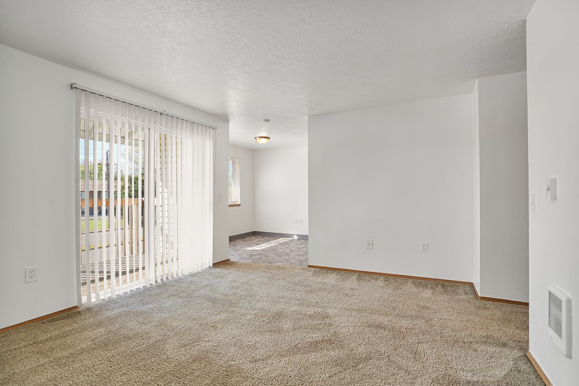 Empty carpeted living room with white walls and sliding glass doors with vertical blinds
