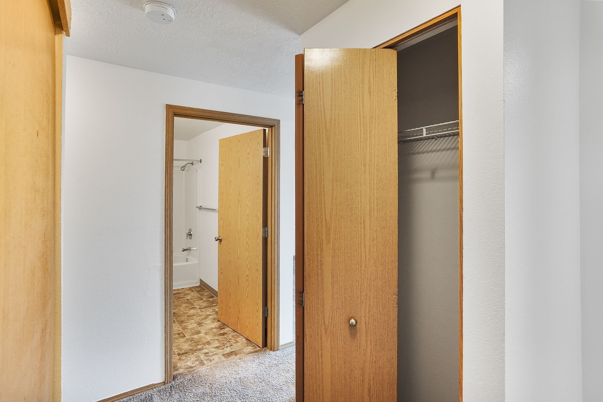 Open closet and doorway in a white bedroom, with a brown door and mirror reflecting a tiled bathroom.