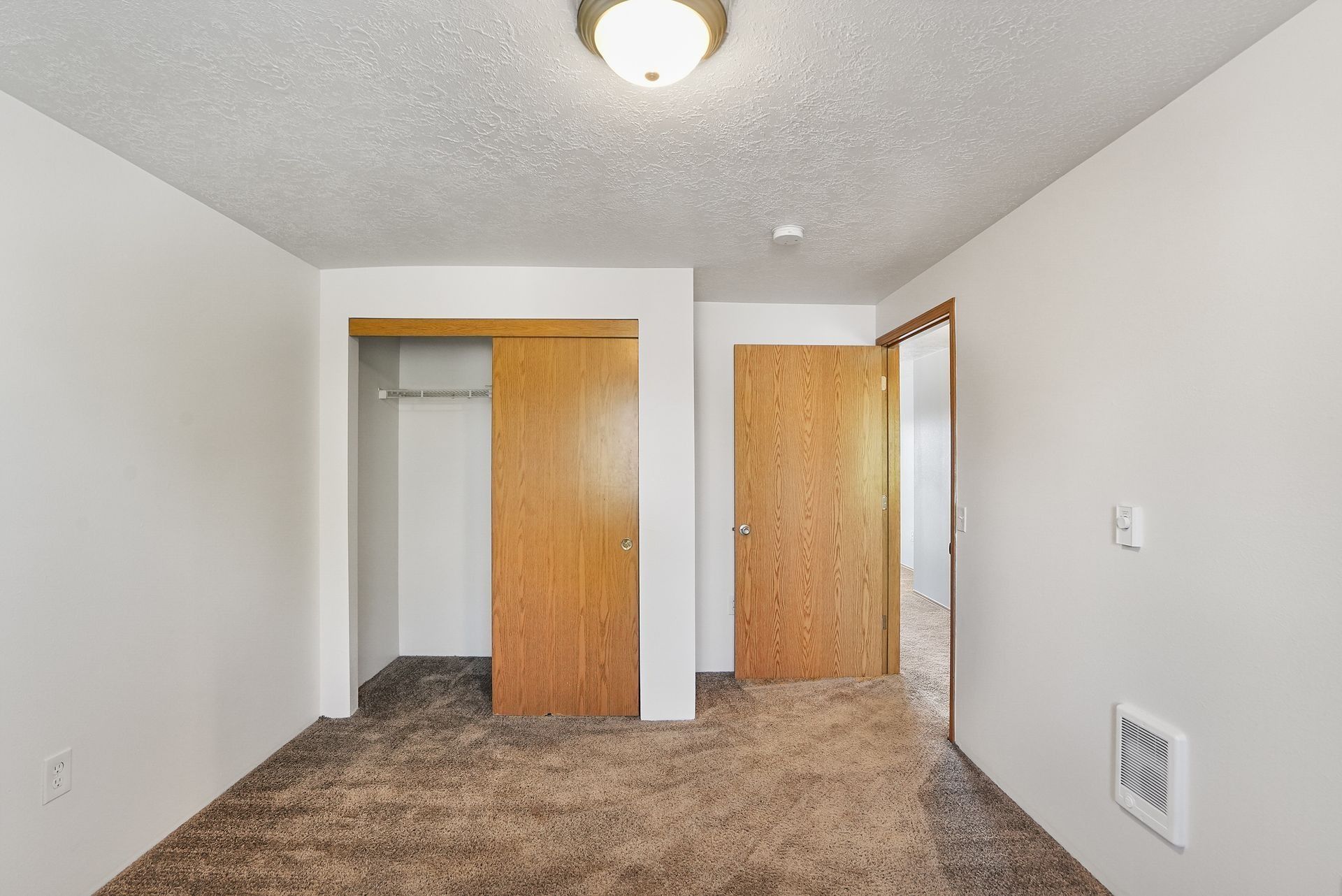 Small empty bedroom with beige carpet, white walls, and two wooden closet doors