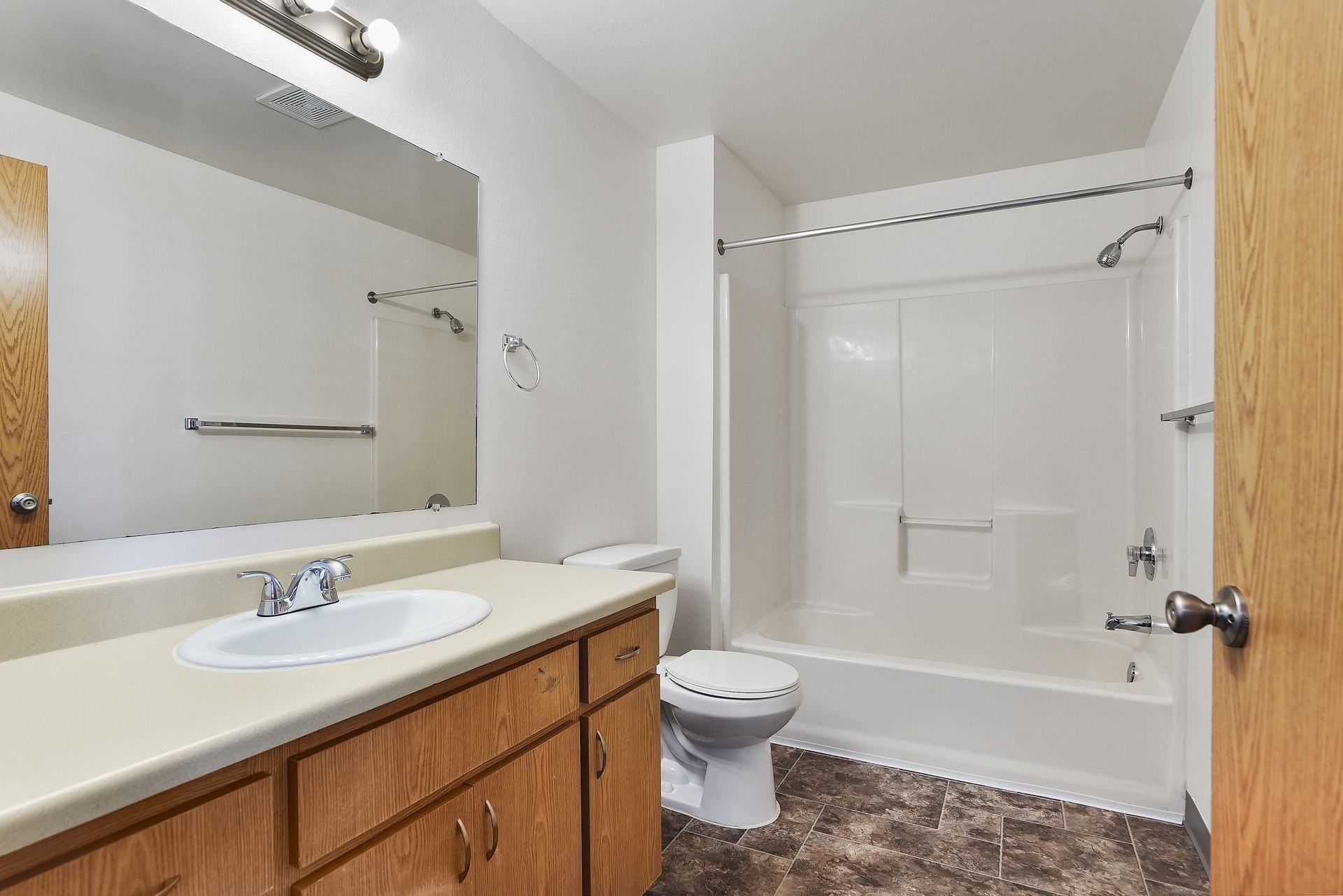 Small bathroom with white tub, toilet, sink vanity, and large mirror above wood cabinets.