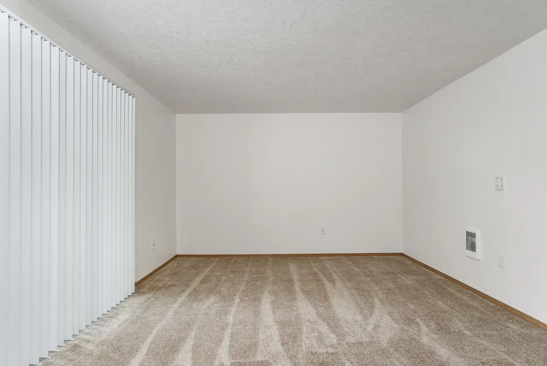 Empty beige-carpeted room with white walls and vertical blinds on the left