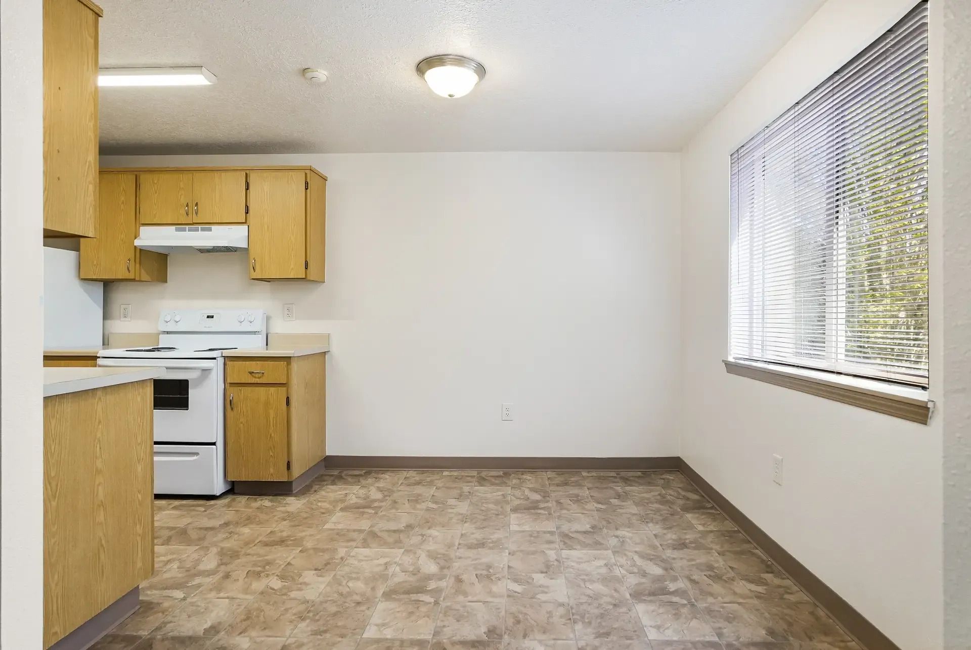 Empty kitchen with tan cabinets, white appliances, and a large window with blinds.