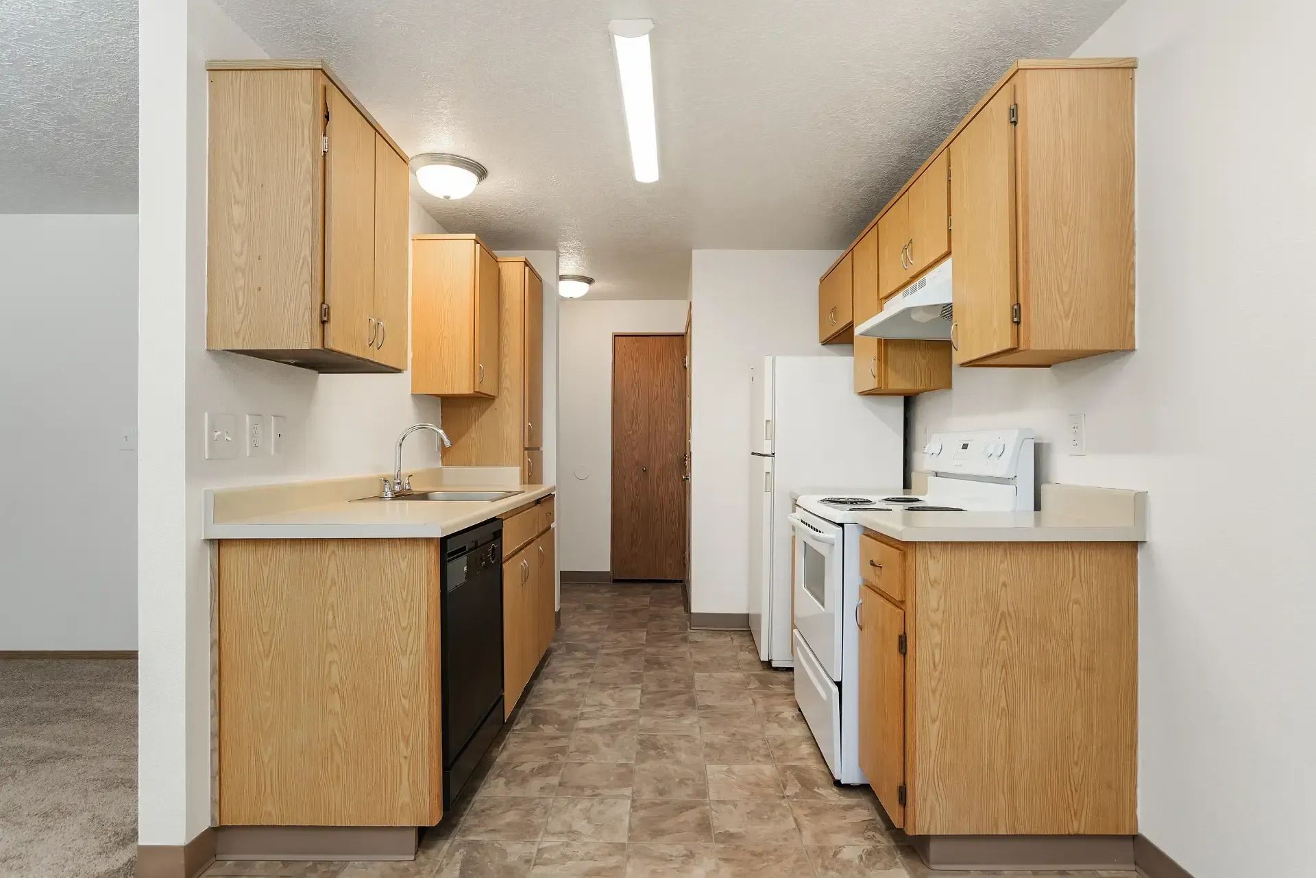 Small kitchen with light wood cabinets, white appliances, and beige flooring in a narrow galley layout